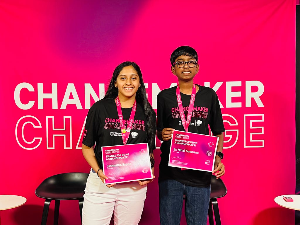 Two young people standing in front of a bright pink background that says “Changemaker Challenge.” They are wearing matching black shirts with the same text on them and holding certificates from a pink lanyard around their neck. The young person on the left is a girl with long black hair and the young person on the right is the boy from the first two photos.