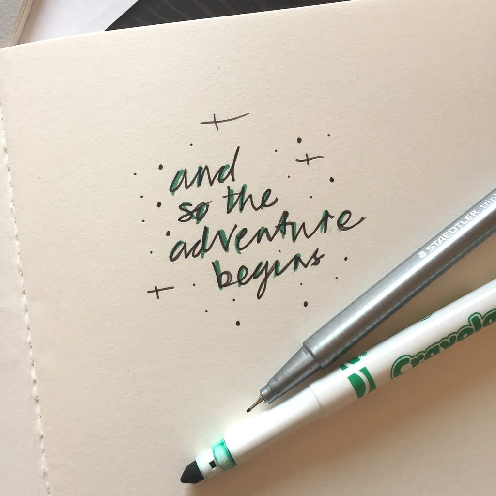 A desk with pens and paper. On the paper, it says, ‘And the adventure begins.’