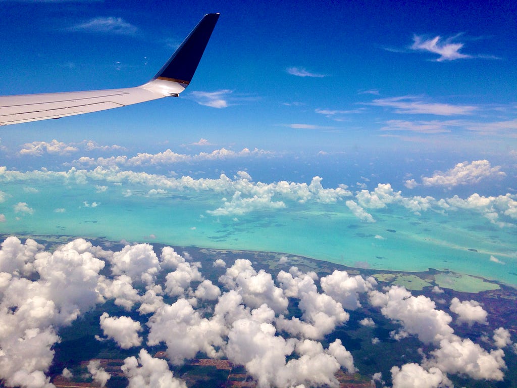 Color photo taken looking out the starboard (right) side of an airplane window, a number of rows behind the wing we can see in the top left of the image, amidst beautiful sunny daylight blue skies, high above the spotted white puffy clouds, with an easy view of the dark land a bit in the foreground, and above turqouise waters featured in the distance as a strip across the snapshot.