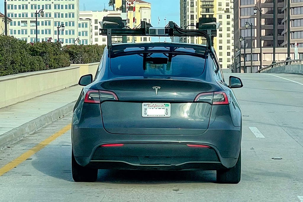 A grey Tesla seen from the rear with a large roof-mounted rack holding a lidar sensor array.