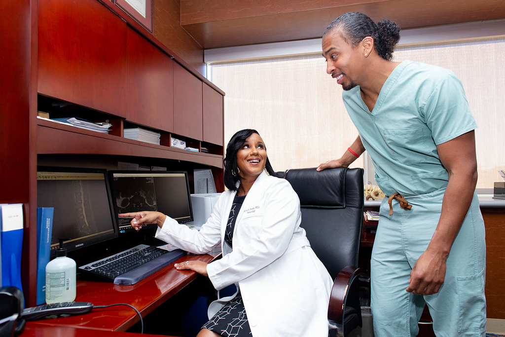 Dr. Porter in a white coat sitting at a desk in a medical office, pointing to a brain scan displayed on a computer monitor. She is smiling and engaged in conversation with a student standing beside her, who is wearing blue-green scrubs. The student is leaning in attentively, looking at the screen. The office includes wooden cabinetry, medical supplies, and multiple monitors.