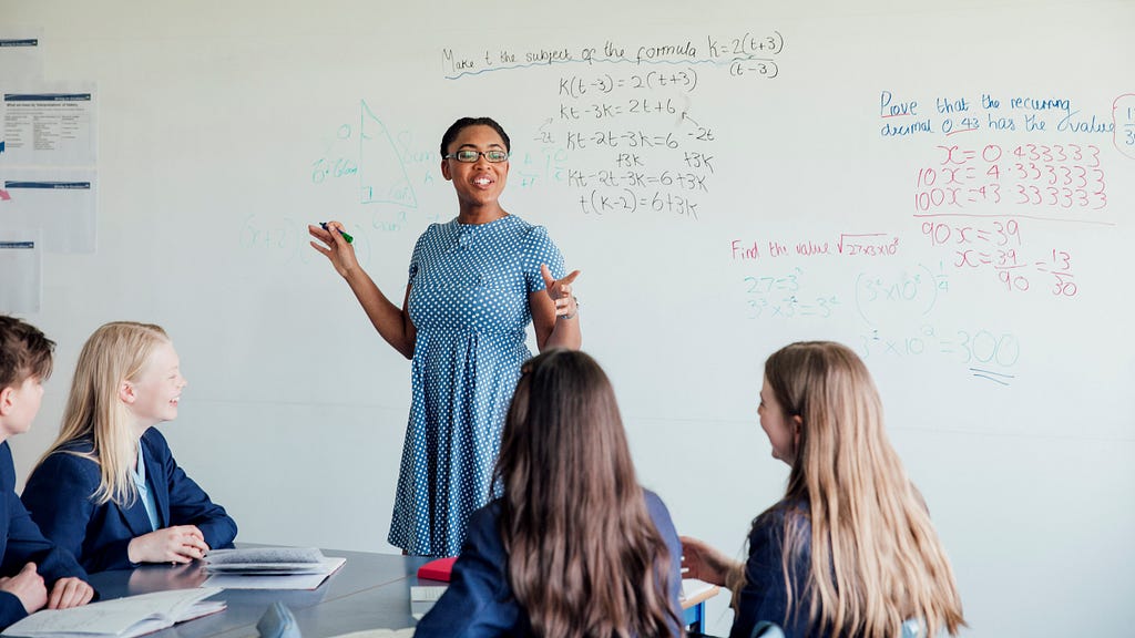 Woman in a light blue polka-dot dress, standing in front of a white board with math equations while 3 female students in school uniforms look forward at her.