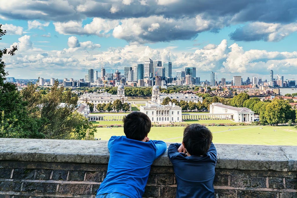 Two boys look out over a wall with the greens of Greenwich Maritime Museum in the foreground and Canary Wharf in the distance