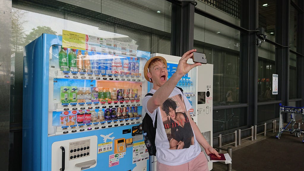 Chris in a Whitney Houston t-shirt, taking a selfie in front of a vending machine at Tokyo Narita Airport.