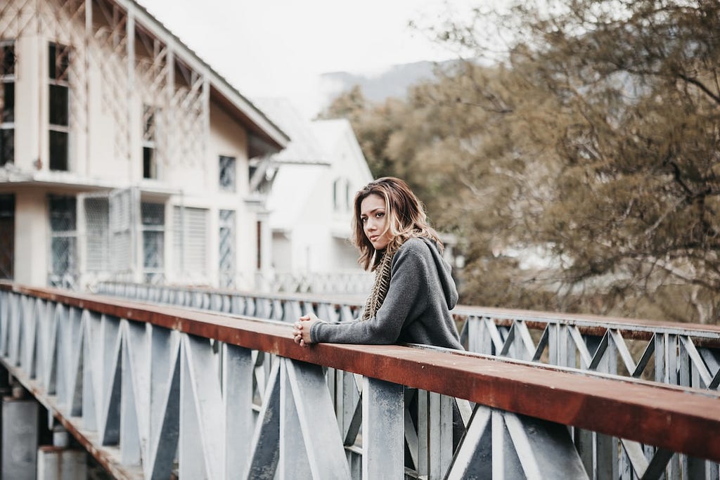 A slender, fair-skinned lady stands on a bridge with her folded hands placed on the metal and wood rails of the bridge asooks on in deep thoughts.