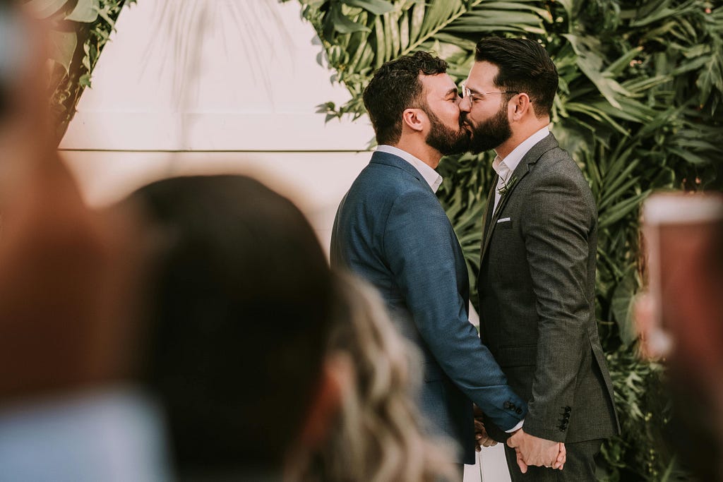 A photo shows two grooms dressed in suits kissing. The grooms are in focus, while the wedding guests who are looking on are shown out of focus and from behind. Behind the grooms is some tropical foliage.