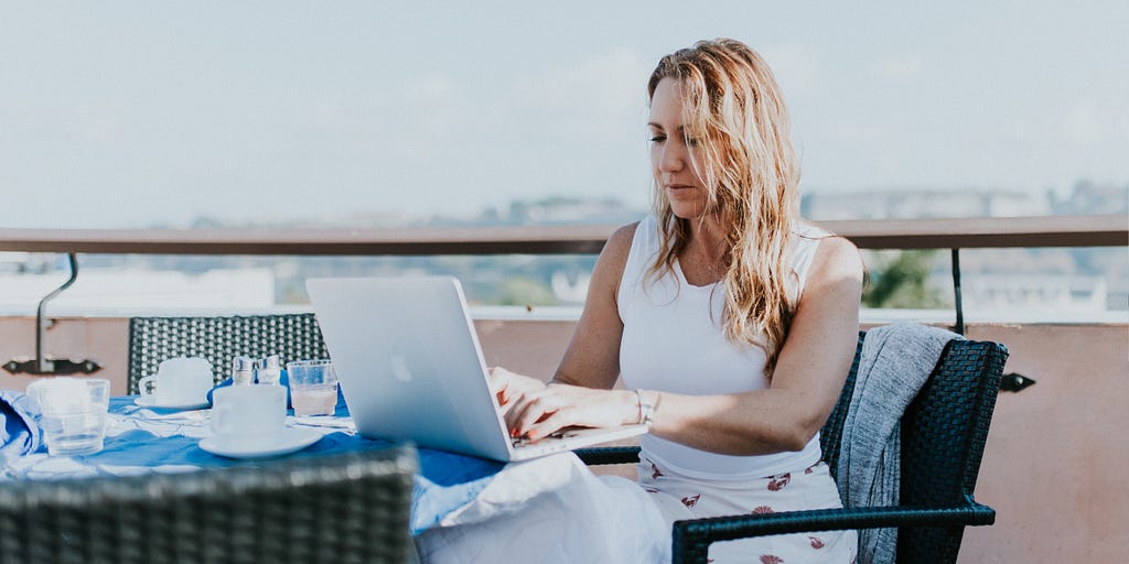 Image of a woman working on her laptop at a table on a balcony overlooking a cityscape