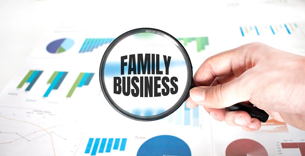 Hand holding a magnifying glass over the words “Family Business” with blurred business charts and graphs in the background.