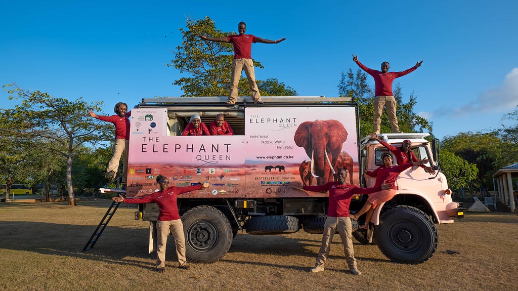 An all-Kenyan outreach team of 9 people pose in front of and inside a 4x4 Bedford Iorry mobile cinema truck decorated with“The Elephant Queen” film images.