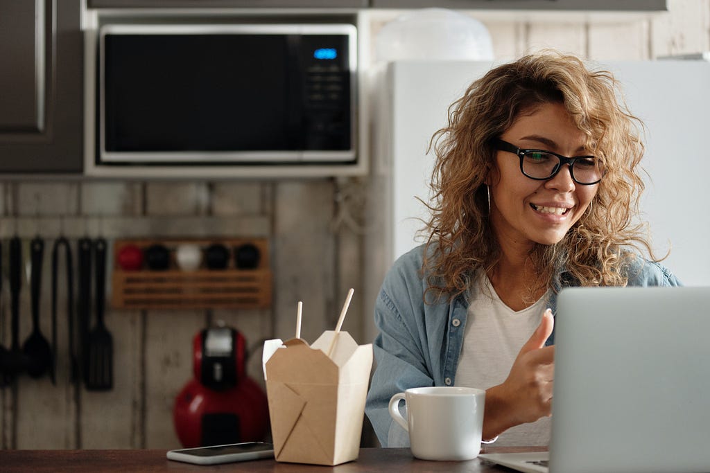 A woman in glasses smiles in front of her computer. An open bento box and coffee mug sit to her right.