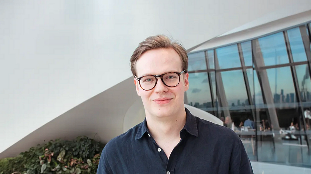 A young man with short, light brown hair and glasses is standing indoors in front of a modern, curved architectural structure. He is wearing a dark blue shirt and is smiling softly at the camera. Behind him, there is a large window reflecting a city skyline in the distance, and some greenery is visible in the foreground.