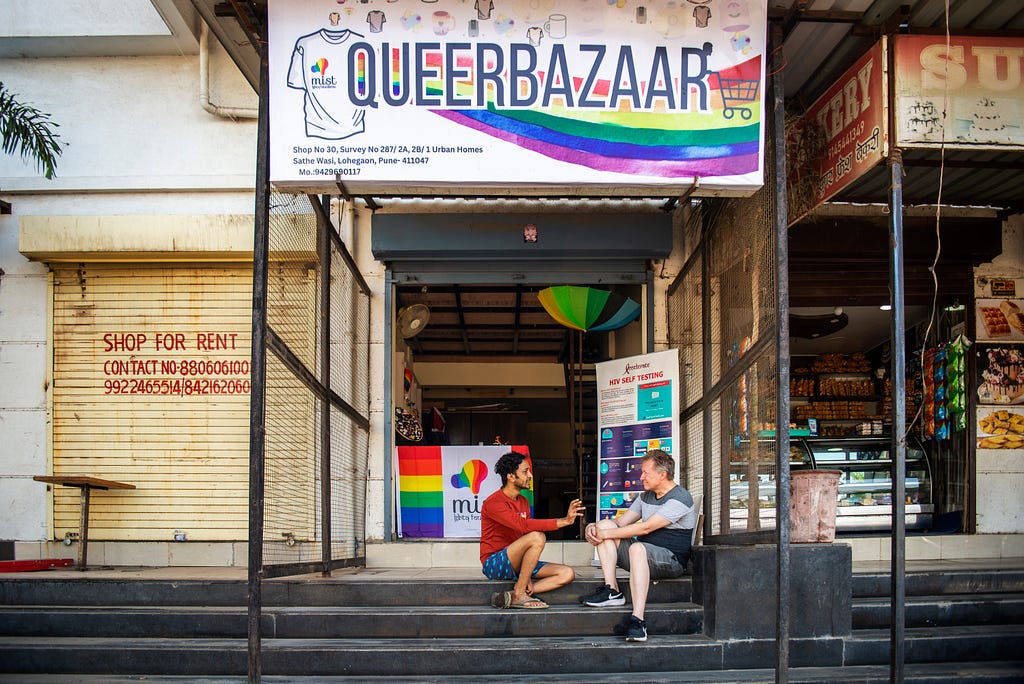 Two men sit conversing on the steps outside of a store. The sign above them says “QueerBazaar.”