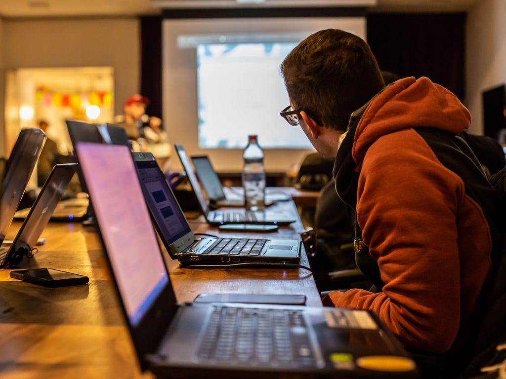 A room with a table that has several laptops on it. Sitting behind the laptops are people who are looking towards a projector screen.