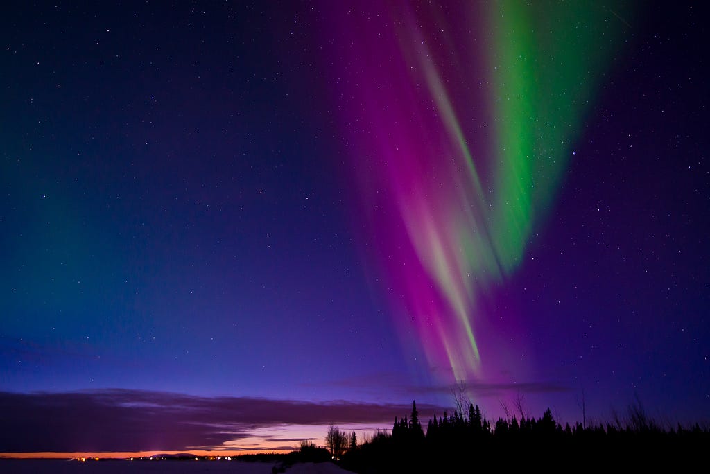 A band of purple and green light waves over black trees and the distant lights of a town.