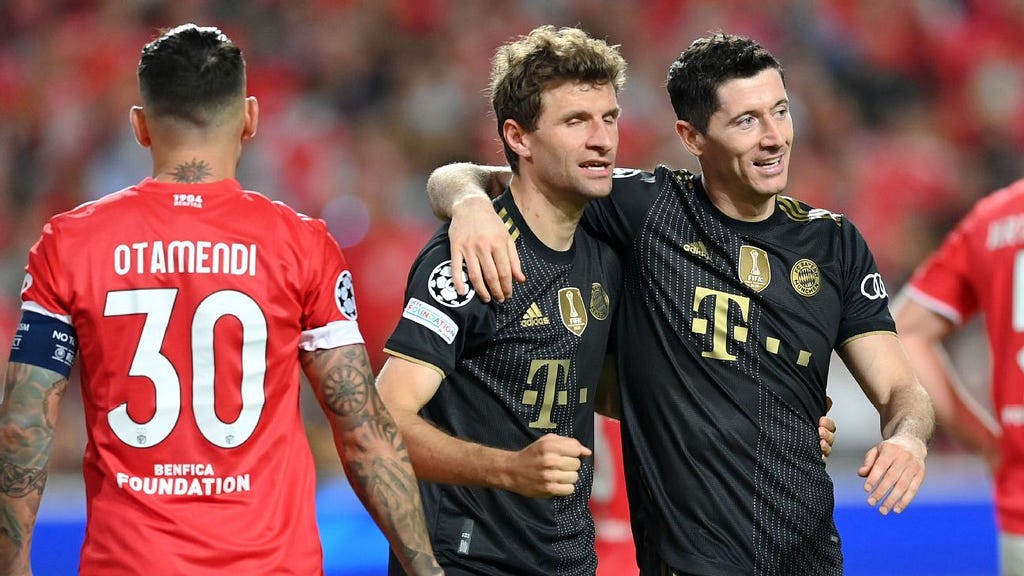 Os jogadores Thomas Müller e Lewandowski comemoram o gol. Eles usam camisa preta com dourada do Bayern de Munique. Ao lado esquerdo e de costas, Otamendi está com a camisa 30 do Benfica.