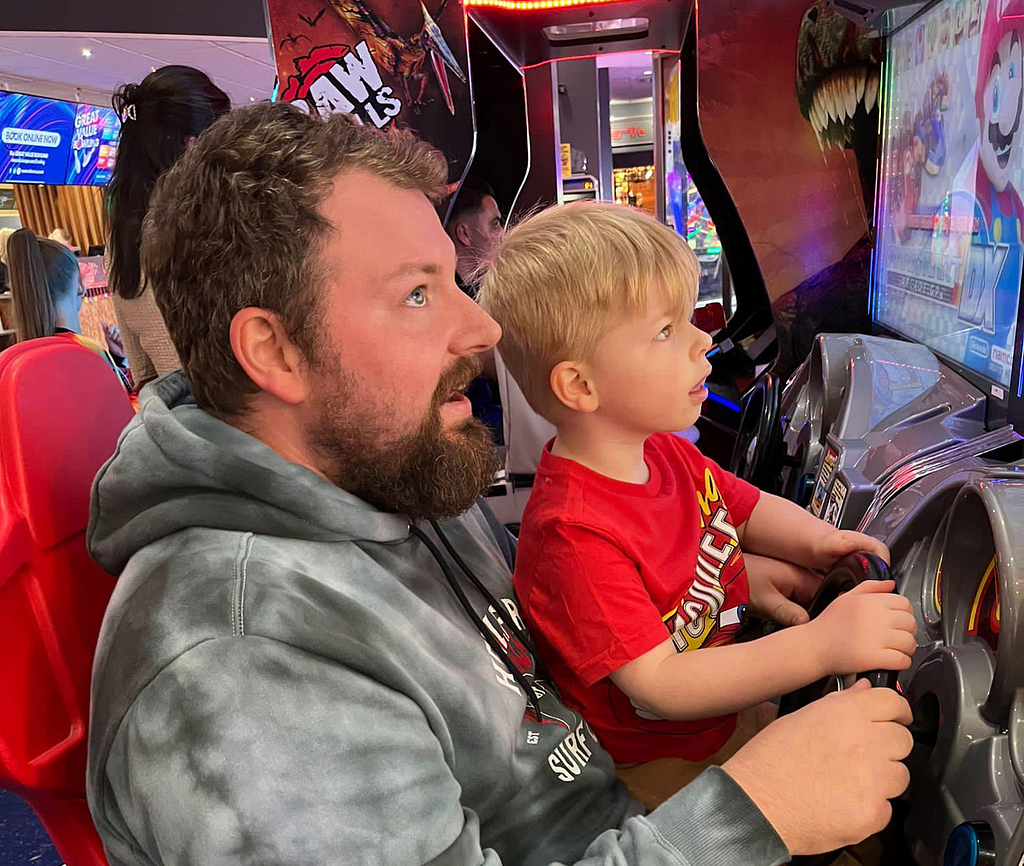 Photo of a bearded Jon and his four year-old son playing a racing game in an amusement arcade. They are staring open-mouthed at the screen.
