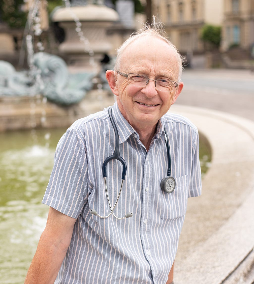 A man with glasses and blue striped shirt with stethoscope around his neck sits on the side of a water fountain, smiling at the camera