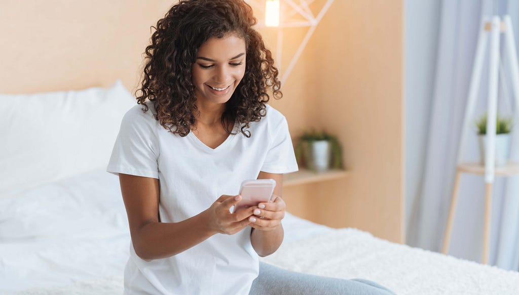 Woman wearing casual clothing sitting on her bed looking at a screen of her smartphone.