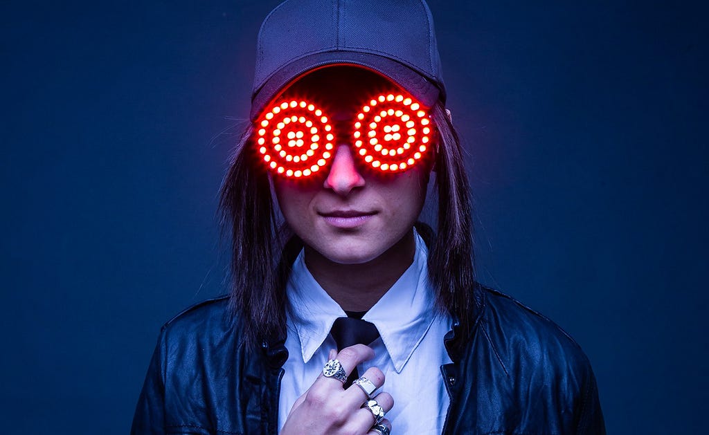 Canadian DJ Rezz posing for the camera wearing LED lit glasses and wearing a trucker hat.