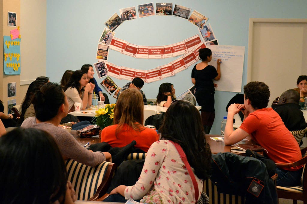 Students are seated at various tables in a room. Up on the wall, the GlobeMed logo is made using pieces of paper with photos and descriptions of GlobeMed values. One person is writing on chart paper on the wall next to the created logo.