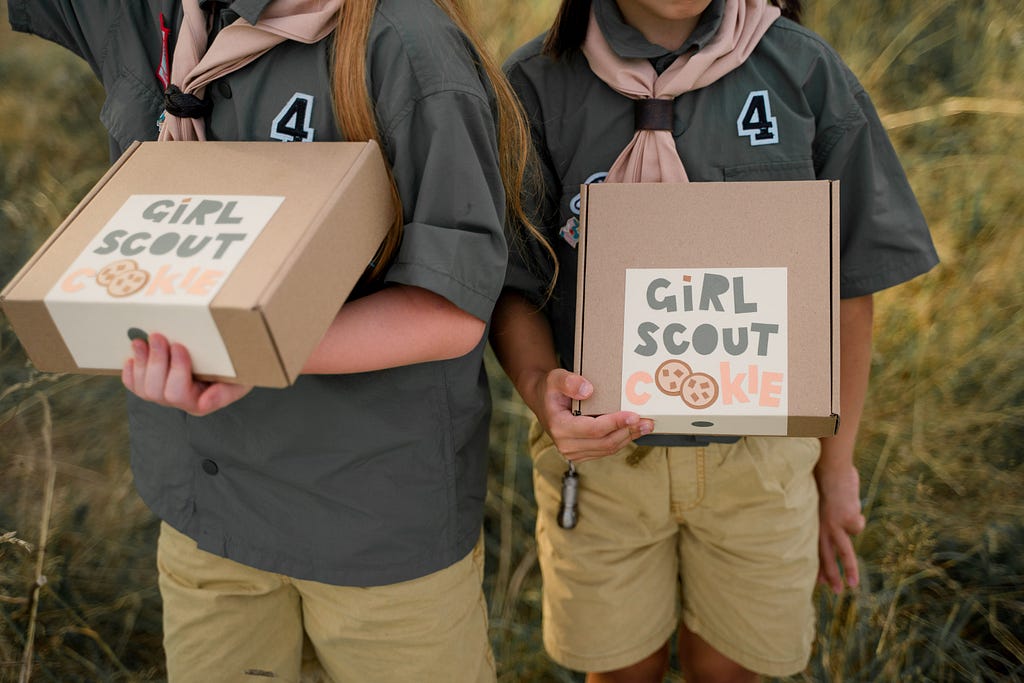 Two girls holding boxes with the words “Girl Scout Cookies” handwritten on them