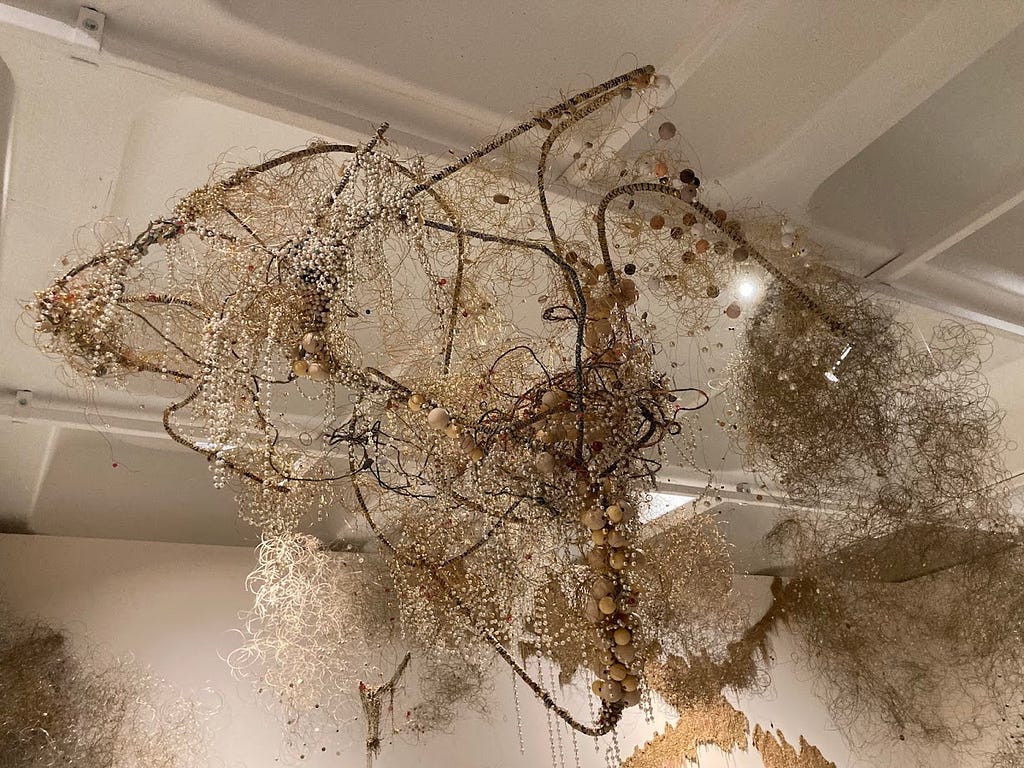 A sculpture of beads and wood, plastic, glass, stone, shells, fabrics, various ropes and twines to create almost a cloud-like entanglement hanging from an art gallery ceiling.