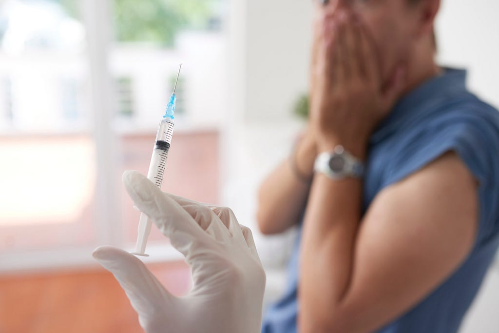 A person holds their hands over their mouth in fear as a medical professional holds up a syringe.