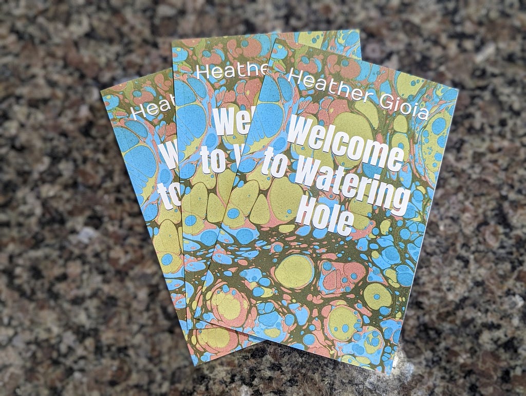 Three copies of “Welcome to Watering Hole” sit on a granite counter top. The book covers are a marbled combination of dark green, light green, light blue, and rust.