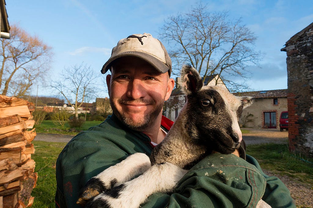 A man in a green jacket and ball cap holding a black and white lamb in a rural setting