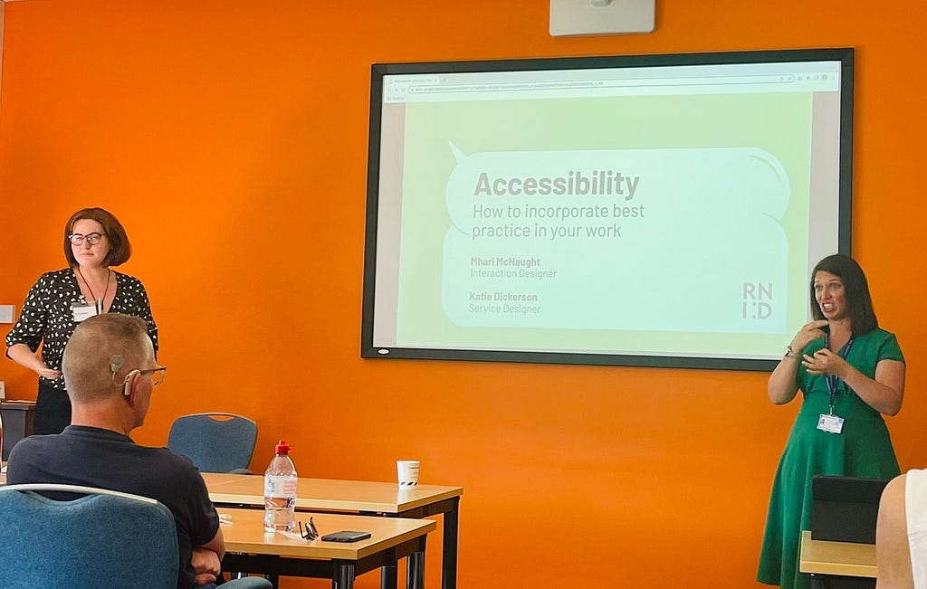 Two women stand on either side of a screen that says ‘Accessibility: how to incorporate best practice in your work. Mhari McNaught, Interaction Designer, Katie Dickerson, Service Designer’. The woman on the right is doing BSL interpretation. There is a man sitting who is sitting in front of a table and looking at the screen who is wearing a hearing aid.