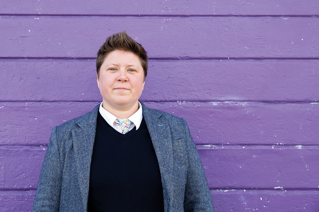 Carrie Bishop standing in front of a purple wall wearing a shirt, jacket and colourful tie