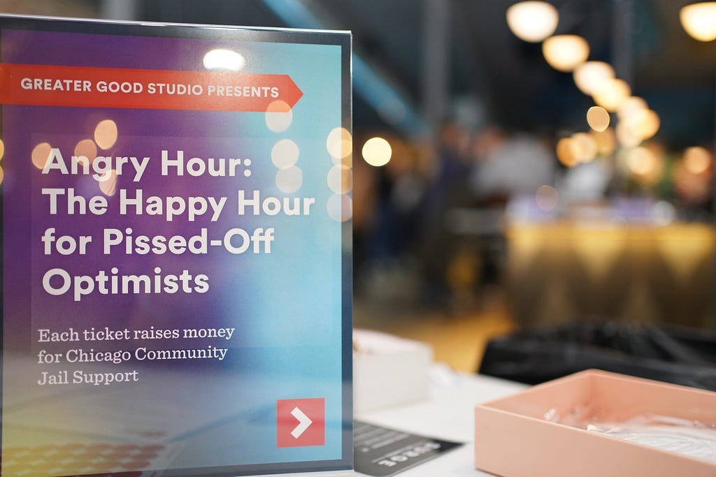 A paper sign in an upright plastic holder. The sign says, “Angry Hour: The Happy Hour for Pissed-Off Optimists.” There’s a blurry bar in the background.