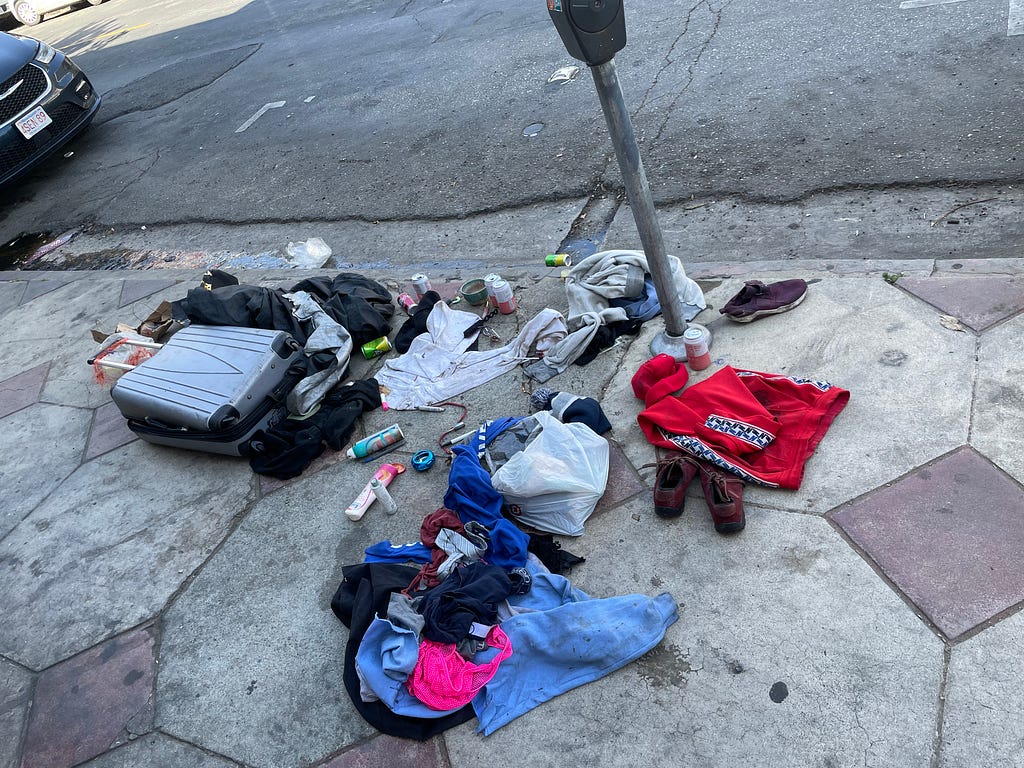 A dirty sidewalk near Hollywood Boulevard in Hollywood California, on it is a broken suitcase and clothes and other personal belongings strewn about the haphazard way. The owner nowhere to be seen…