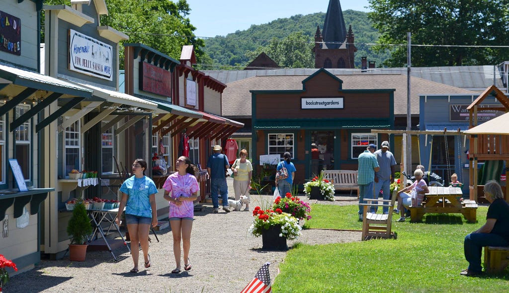 A row of little sheds with 1880s style false fronts holds micro-businesses. Customers are strolling, shopping, talking to each other and sitting on benches in the grassy open space.