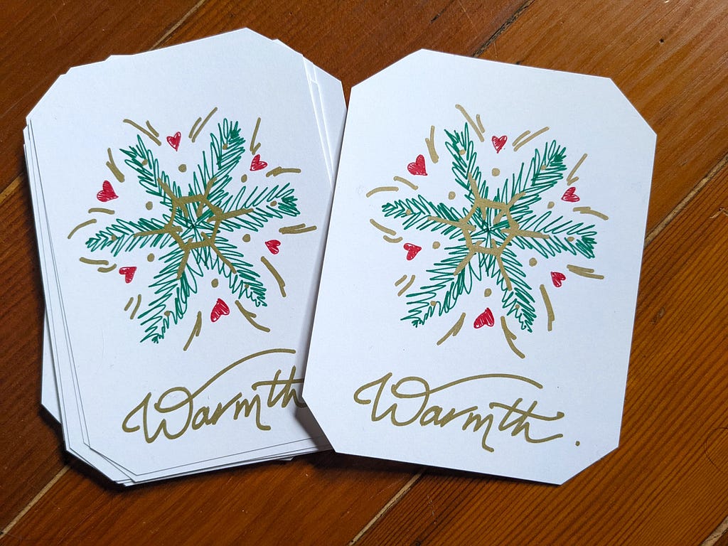 Two white cards laying on brown floorboards, with their corners cut off; the left one sits atop a small stack. The paper shows a “snowflake” design, made of green fir sprigs, red hearts, and gold embellishments. Underneath is “Warmth.” written in gold script.