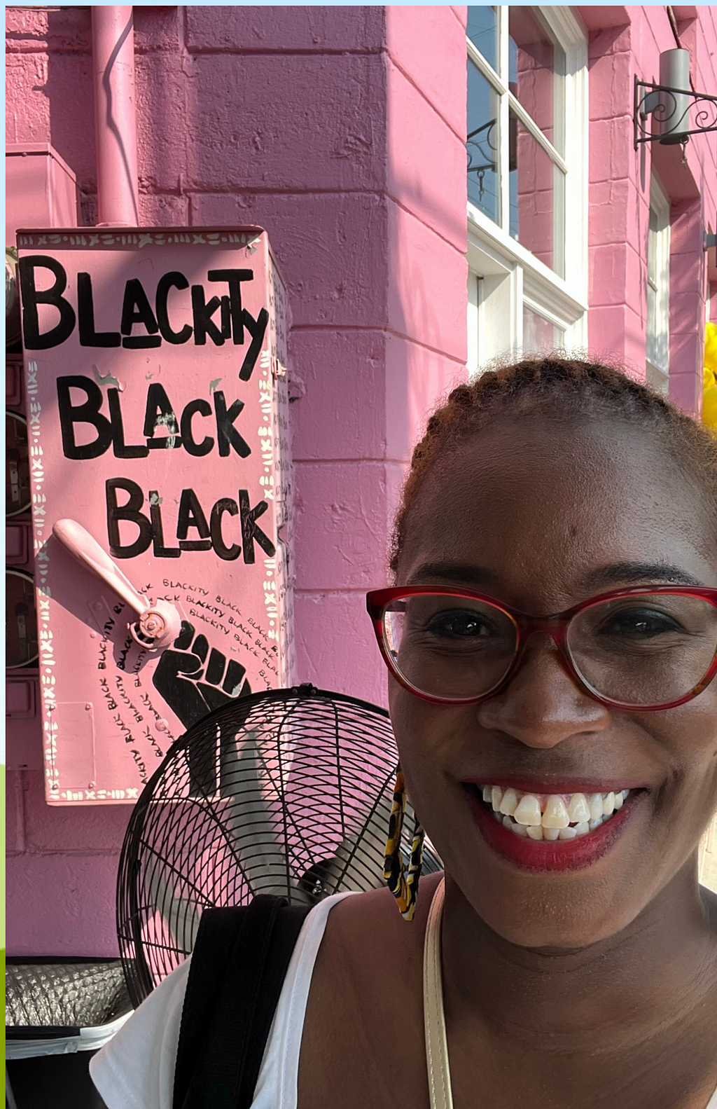 Black woman smiling in front of pink sign with black letters reading “Blackity Black Black”