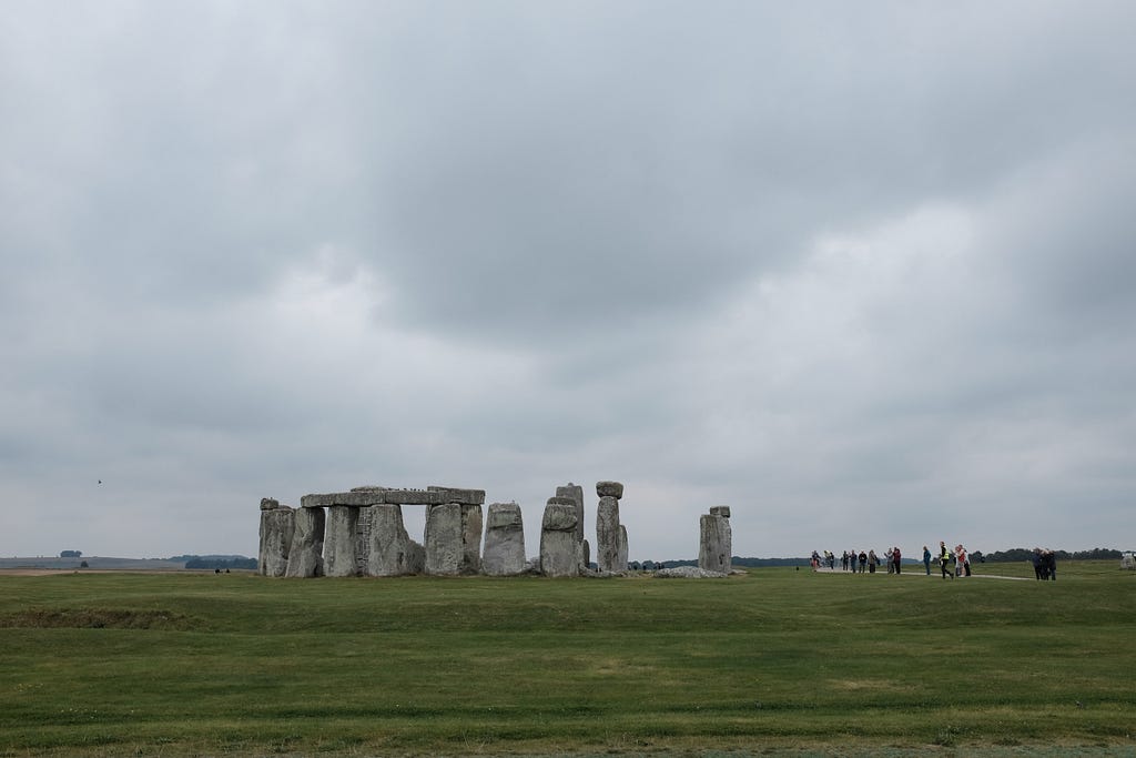 Stonehenge from a distance with a group of people to the right. Grey cloudy skies