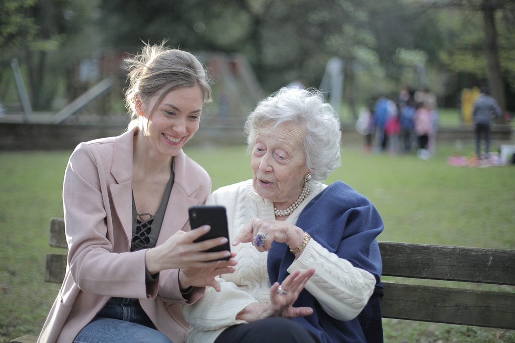 Cheerful senior mother and adult daughter sit on a bench in a park enjoying something on the daughters phone together.