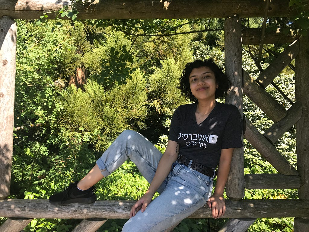 Person wearing black shirt with Hebrew letters and blue jeans sits on wooden horizontal pole with one leg up and one down.