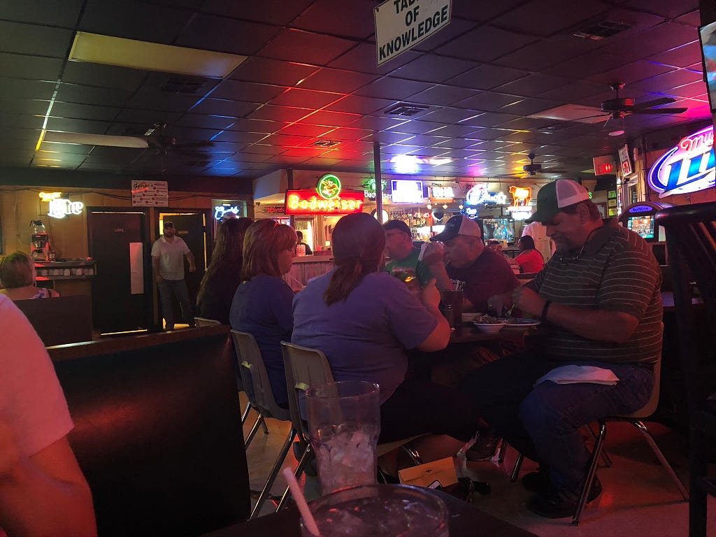 A group of six people are seated at a table in a sports bar full of neon advertisements for cheap beer (Budweiser, Miller Lite, etc.) Two are wearing matching ball caps.