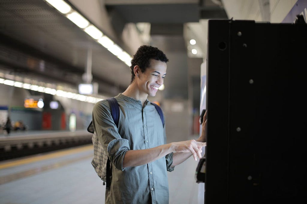 Een glimlachende jongeman koopt tickets aan een automaat op het perron van zijn metro of trein.