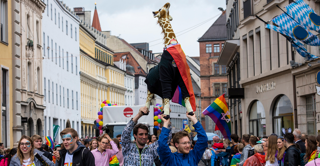 Picture of the pride parade in munich, people holding a large giraffe stuffed animal in the air celebrating pride.