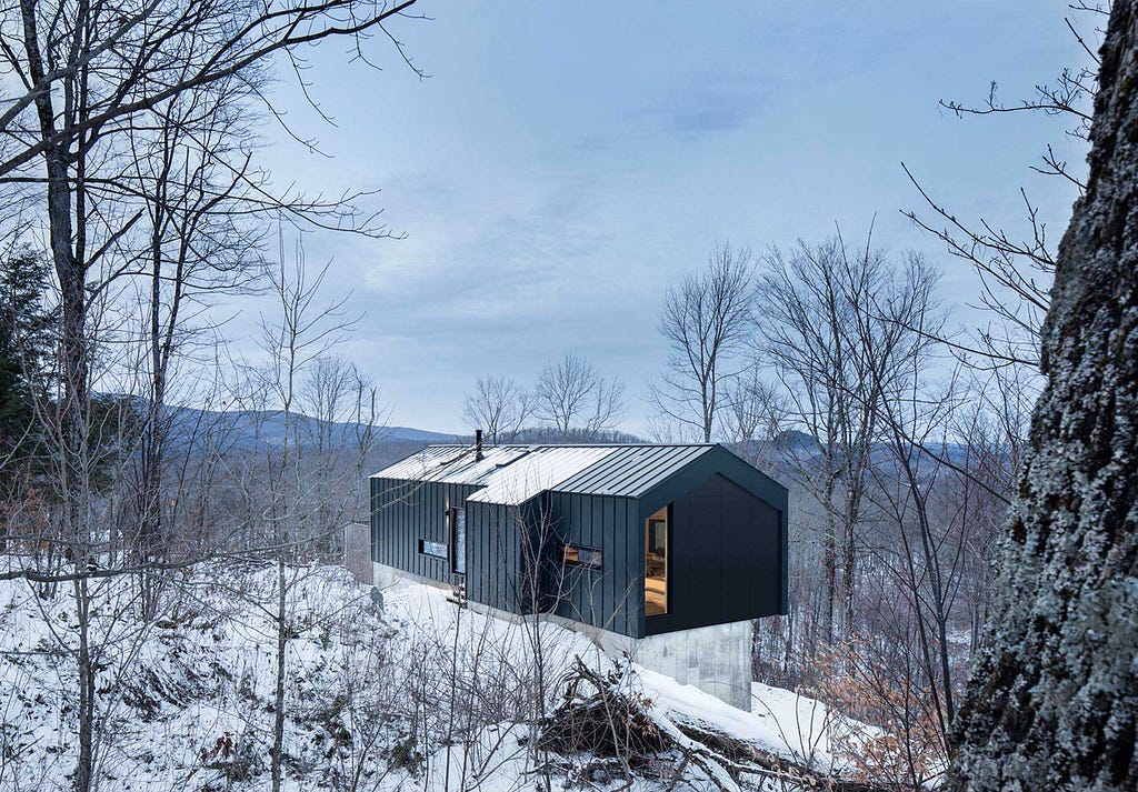A two storey, black and wooden-clad building sits on a natural plataeu surrounded by snow.