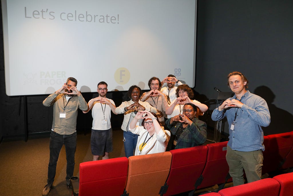 Nine people, making heart shapes with their hands, are grouped in front of a cinema screen that has the words: “Let’s celebrate!”