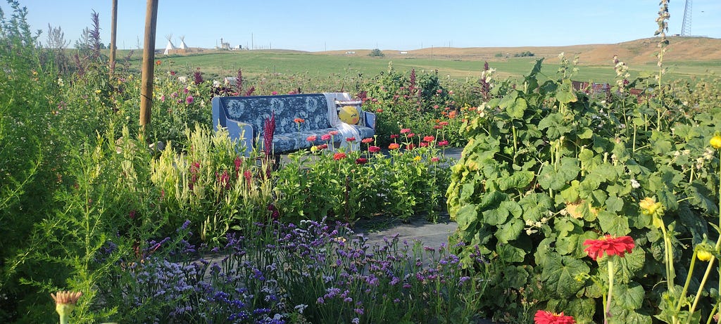A blue and white couch in the middle of a flower garden which a white blanket and yellow pillow.