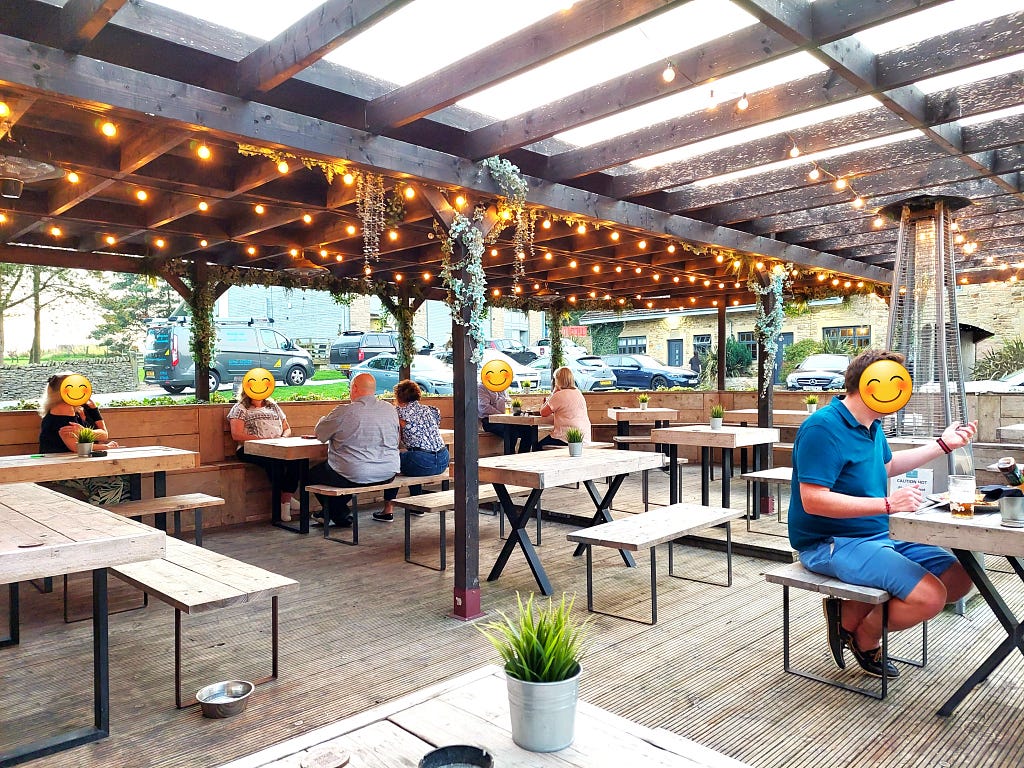 a wooden deck with wooden tables and chairs. A few people are sat around under a pergola roof, hung with fairy lights.