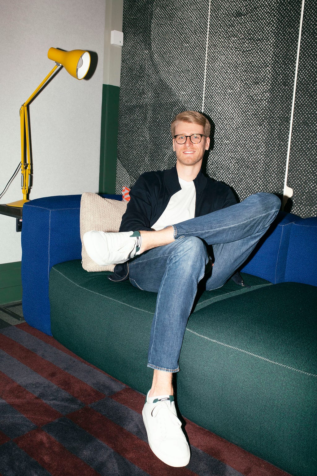 A blond man wearing glasses sitting on a green couch and smiling