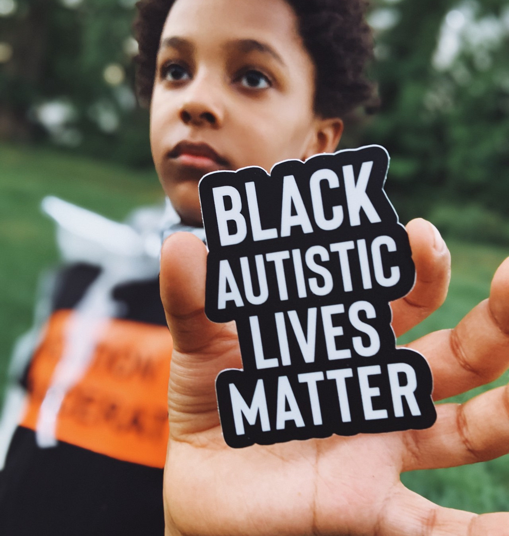 A Black child outdoors holding a sticker that reads “Black Autistic Lives Matter”