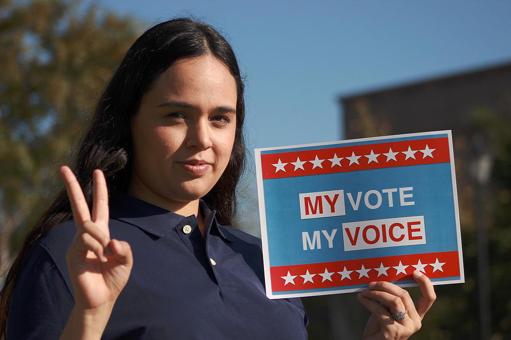 A young Latina woman makes a peace sign with her right hand and holds a sign in her land hand that reads “My Vote, My Voice.”