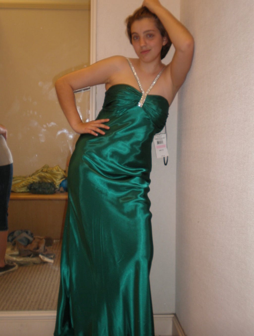 A young white teenage girl strikes a post in an emerald green floor-length gown in a dressing room. She has short brown hair and one of her hands is on her hip while the other is resting against the wall. A tag is hanging from the dress and a mirror is visible in the background.